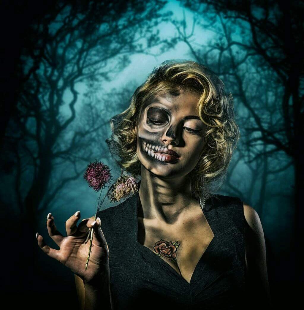 The image shows a woman with skull makeup, holding a flower, with a dark, foggy forest background.