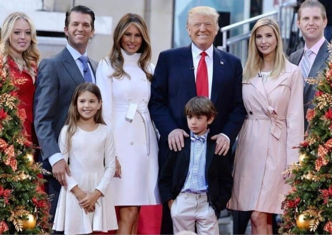 A group of eight people posing for a formal photo on steps outdoors. A man in a dark suit and red tie stands in center, flanked by adults and children.