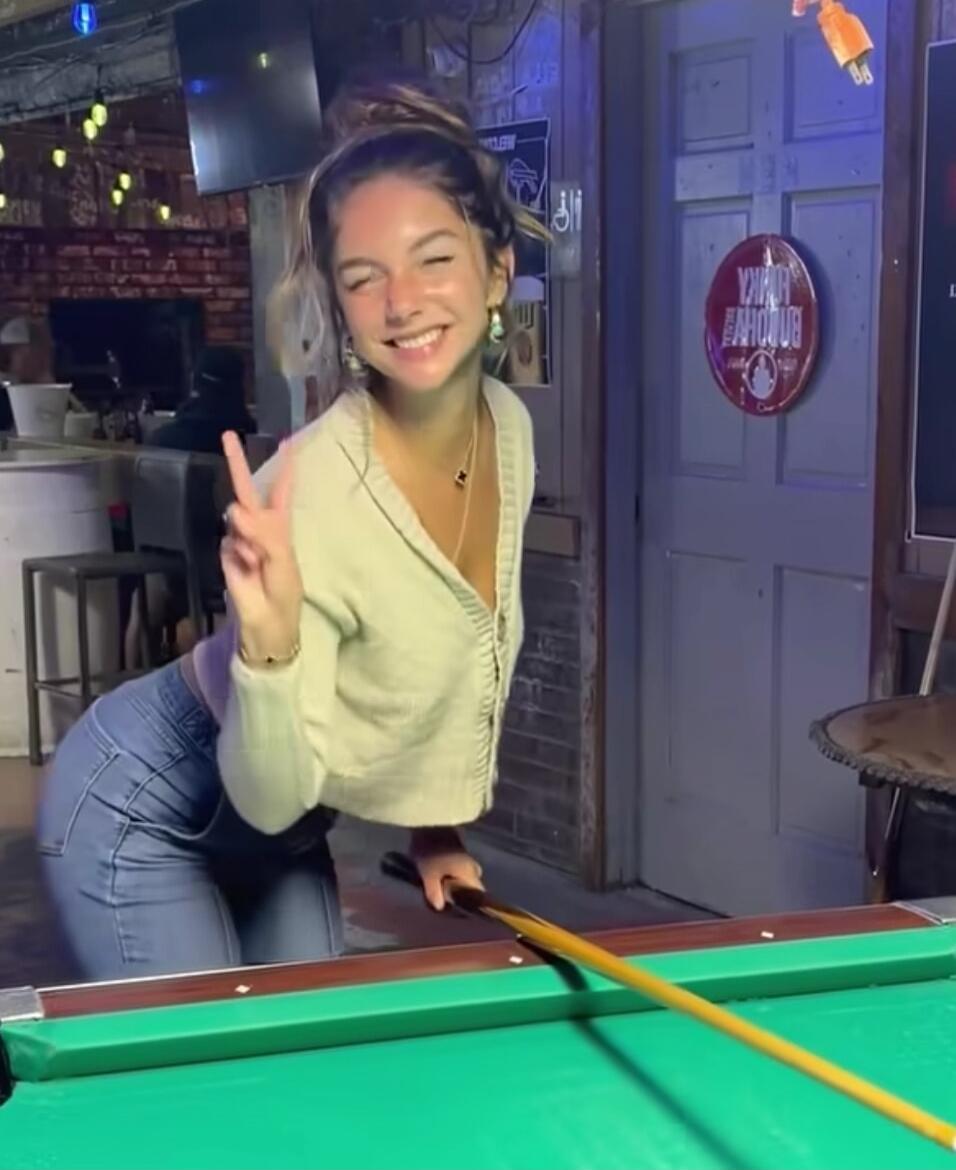 A young woman smiling and making a peace sign while holding a pool cue next to a pool table in a bar setting.