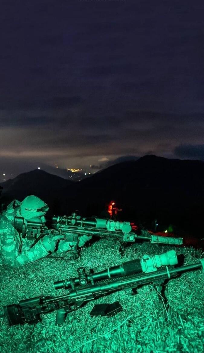 Nighttime military snipers with rifles on a hillside, city lights in distance.