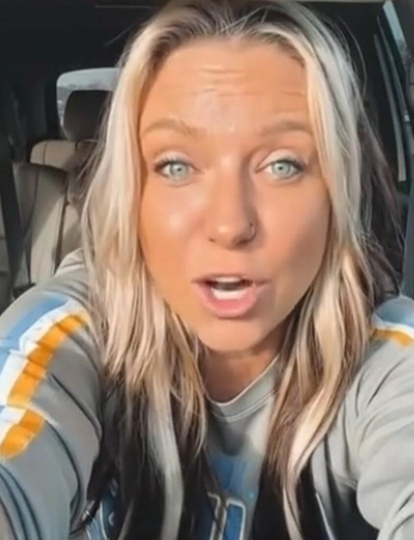 A woman with blonde hair is speaking to the camera from inside a car.