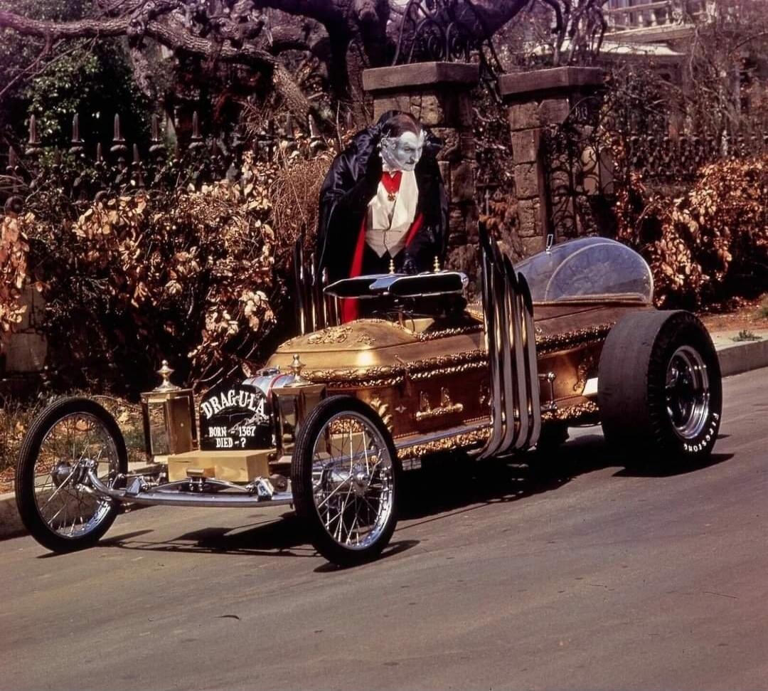 DRAG-ULA BORN - 1367 DIED - ? A man dressed as Dracula stands next to a custom hot rod car designed to look like a golden coffin. The car features large exhaust pipes and a bubble canopy.