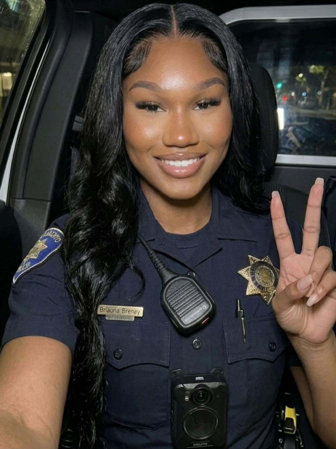 A smiling woman in a police uniform, wearing a name tag that reads 'Briauna Brenay', a radio, and a body camera. She is making a peace sign with her hand. The uniform has a 'Palo Alto' patch.