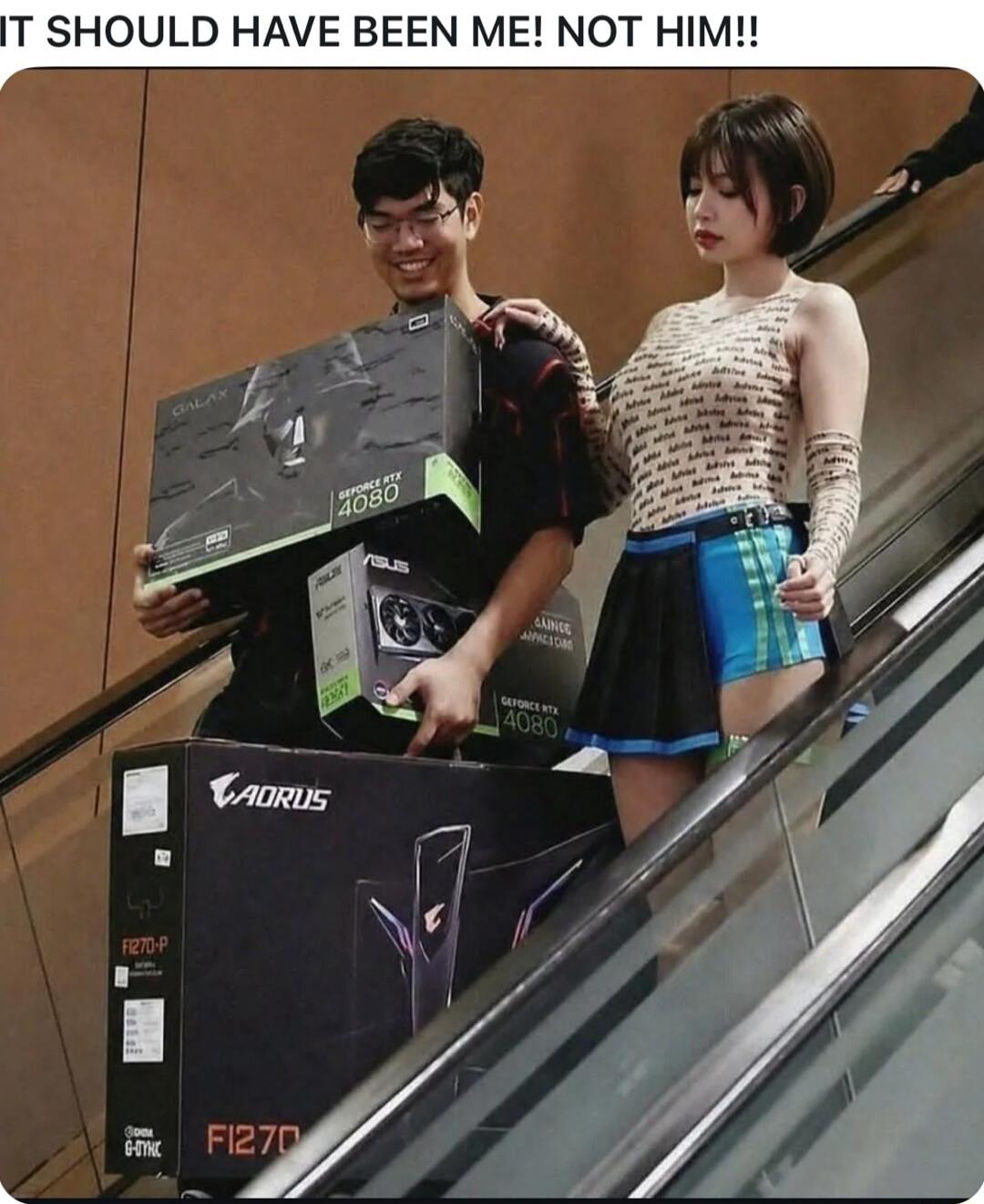 IT SHOULD HAVE BEEN ME! NOT HIM!! A man on an escalator is happily carrying multiple large boxes of computer hardware, including a GALAX GEFORCE RTX 4080 graphics card and an AORUS F1270-P monitor. A woman next to him has a serious expression, with her hand on his shoulder.