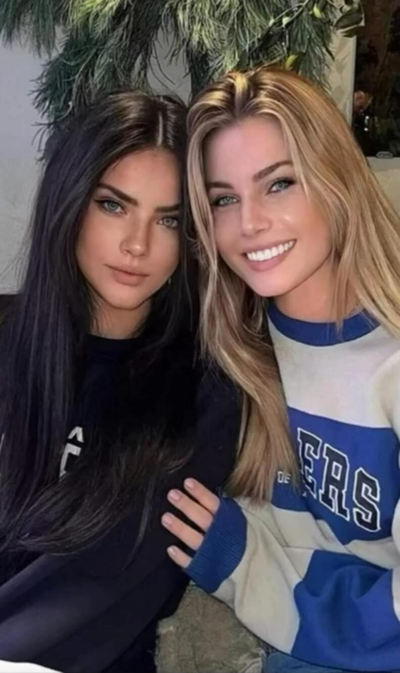 Two young women posing together, one with dark hair and one with blonde hair, both looking at the camera. The blonde woman is smiling.