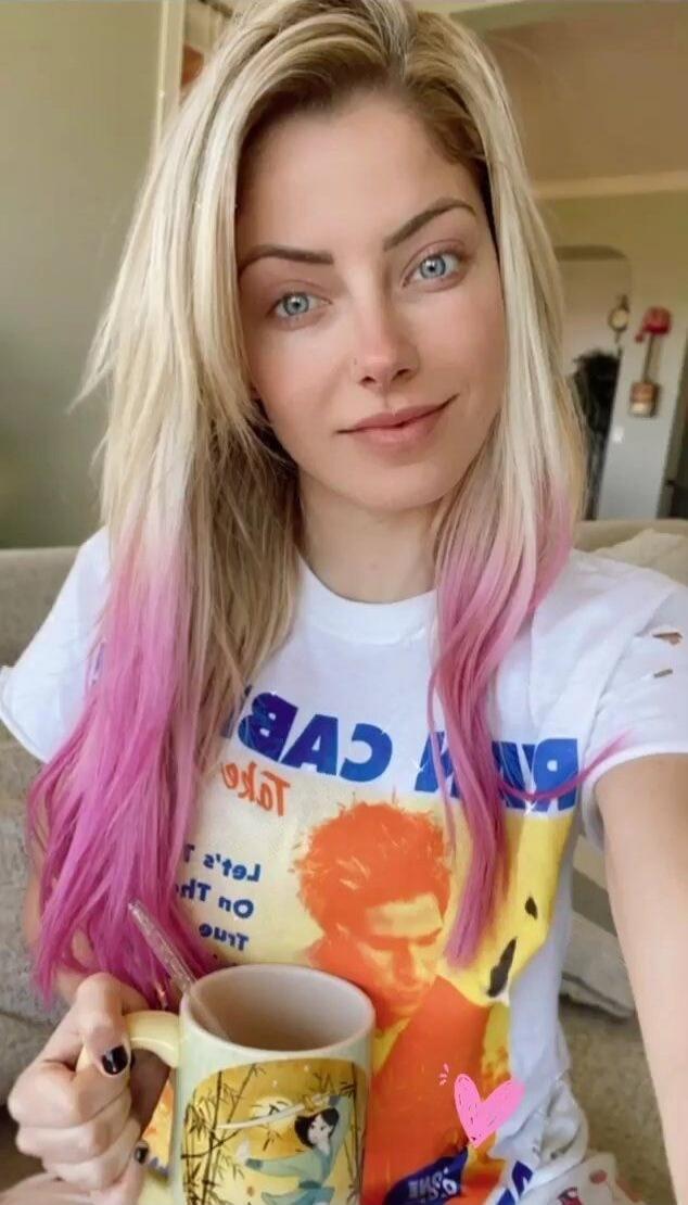 A woman with blonde hair and pink tips, blue eyes, holding a mug. She is wearing a white t-shirt with a graphic design and some reversed text, including 'CVB', 'T'let's', 'HT no', 'ourT'. There is a small pink heart sticker on the image.