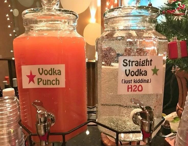 Two beverage dispensers are shown. One is filled with an orange liquid and labeled 'Vodka Punch'. The other is filled with a clear liquid and labeled 'Straight Vodka (just kidding) H2O'. The setting appears to be a party or gathering, possibly during the Christmas season, with festive decorations in the background.