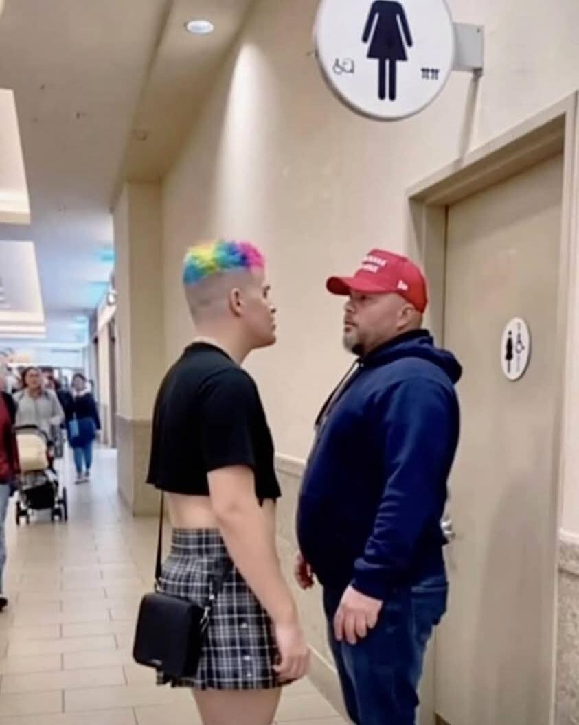 Two individuals are standing face-to-face in a hallway, seemingly in a confrontation. One person has rainbow-colored hair, wears a crop top and a plaid skirt. The other is a man wearing a red 'MAGA' hat and a blue hoodie. A women's restroom sign is visible in the background.