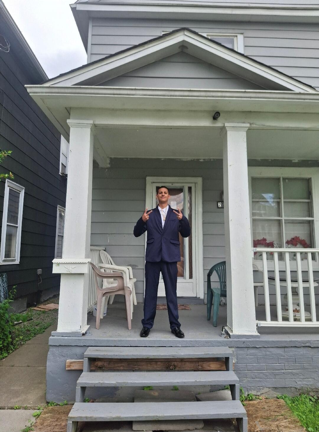 A young man in a dark suit stands on the porch of a grey house, making peace signs with both hands. The house number '28' is visible on the door frame.