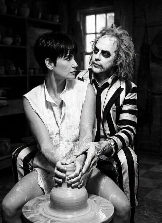 A black and white image showing a man and a woman at a pottery wheel. The man, dressed in a striped suit and with distinctive makeup resembling Beetlejuice, is sitting behind the woman, embracing her and guiding her hands as they shape clay on the wheel. The woman, with short dark hair, looks intently at the clay. The scene is a clear parody of the