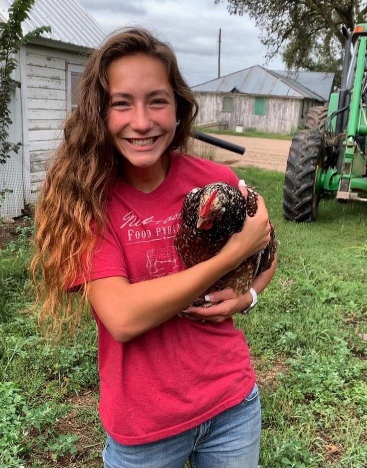 A young woman with long brown hair is smiling and holding a chicken. Her red t-shirt has 'Nebraska FOOD PYRAMID' text on it. In the background, there are farm buildings and a green tractor.