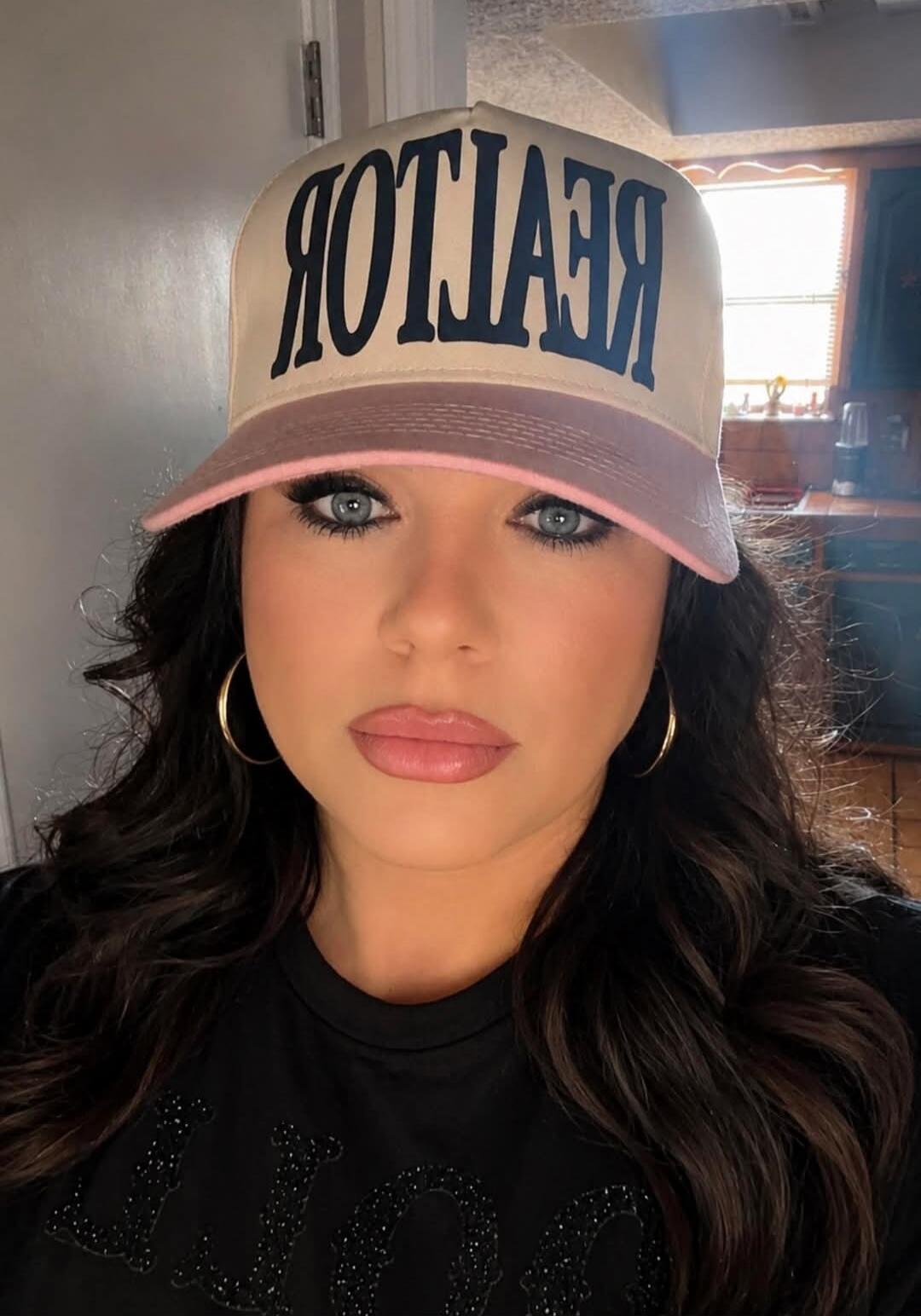 A woman wearing a baseball cap with 'REALTOR' written on it (mirrored). She has dark hair, blue eyes, and makeup, and is wearing hoop earrings.