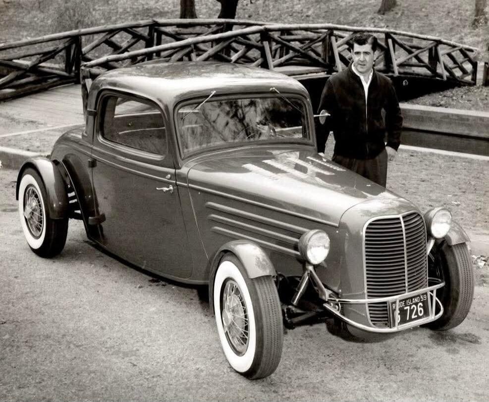A man stands next to a vintage hot rod car. The car has a license plate that reads 'RHODE ISLAND 59' and '726'. A wooden bridge is visible in the background.