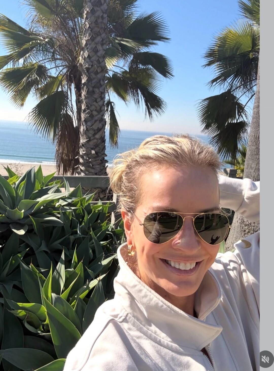A woman taking a sunny selfie with palm trees and the ocean in the background.