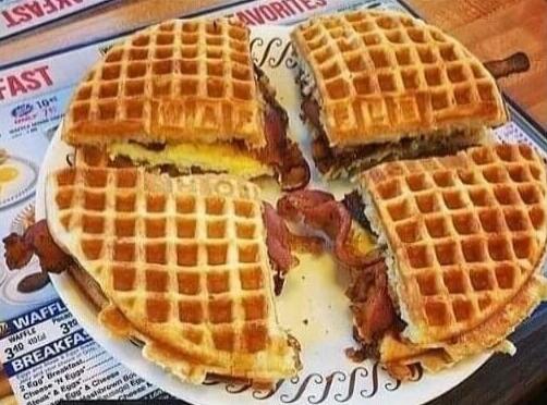 Four square waffles arranged as a sandwich with bacon and cheese/eggs on a plate. In the background, a menu or newspaper shows words like 'FAST' and 'BREAKFAST'.