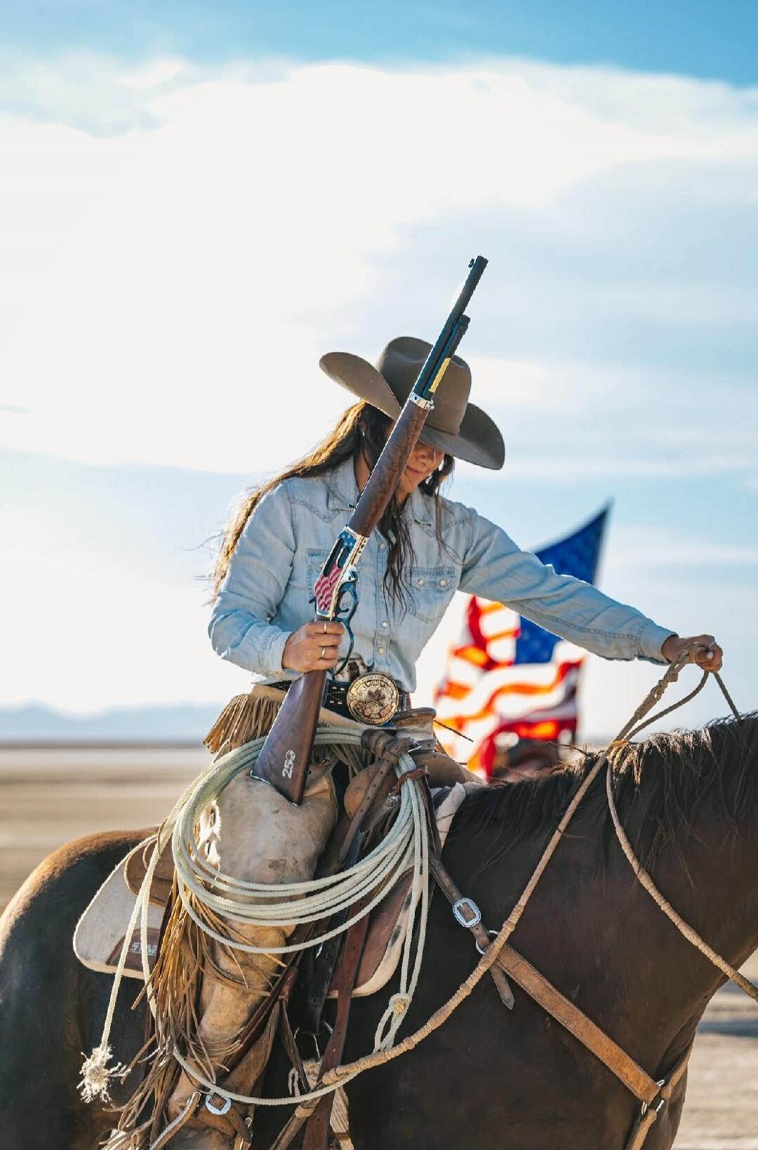 A woman in a cowboy hat and denim shirt is riding a horse, holding a rifle decorated with an American flag design. The number '250' is visible on the rifle stock. An American flag is in the background.