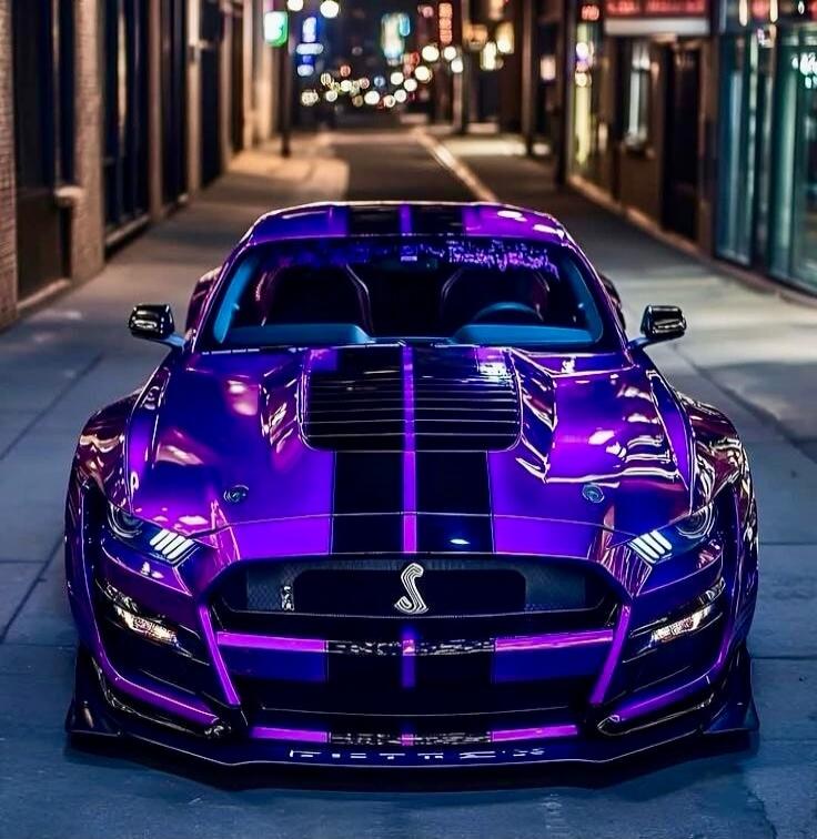 Purple sports car with black racing stripes parked on a city street at night.