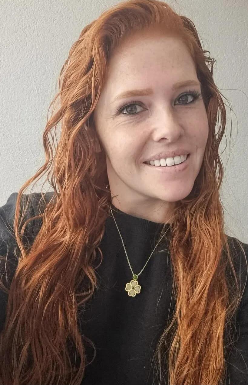 A woman with long wavy red hair, wearing a black top, smiling, and a small four-leaf clover pendant necklace.