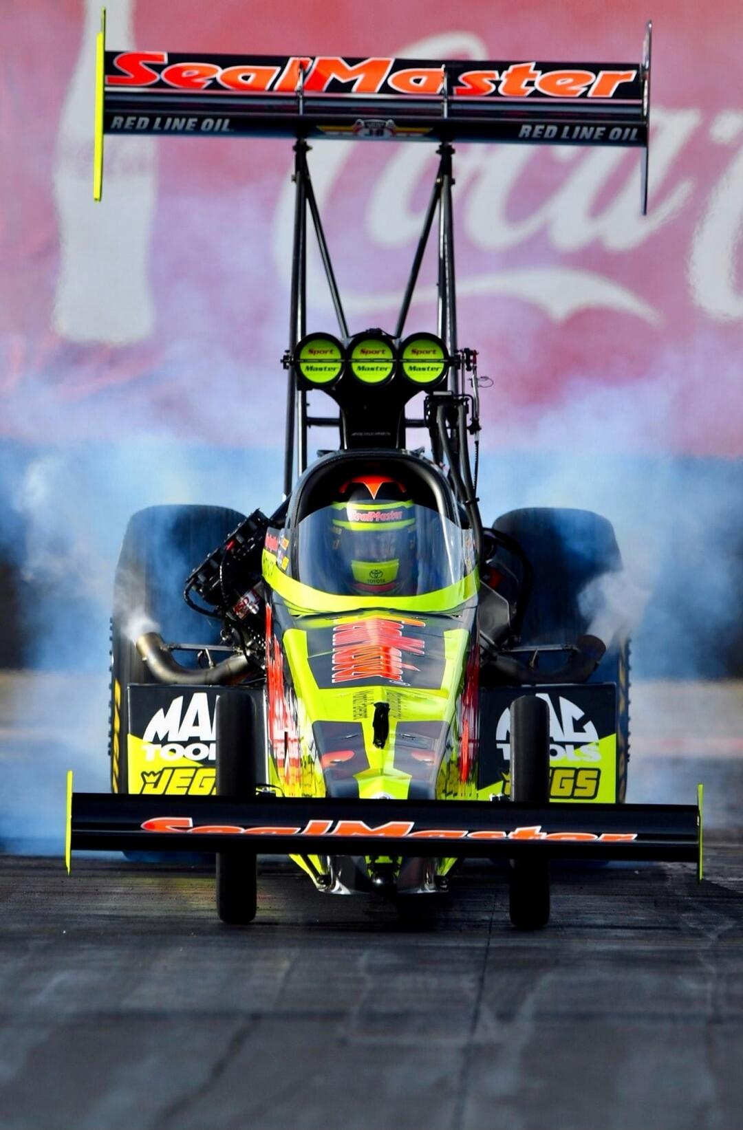 A powerful dragster with 'SealMaster' and 'RED LINE OIL' branding on its rear wing, and 'MA TOOLS' and 'GS' on its side, performs a burnout on a race track, emitting smoke from its tires. The car is brightly colored in neon yellow, black, and red, with 'Sport' and 'Master' visible on its lights and 'Toyota' on the cockpit.