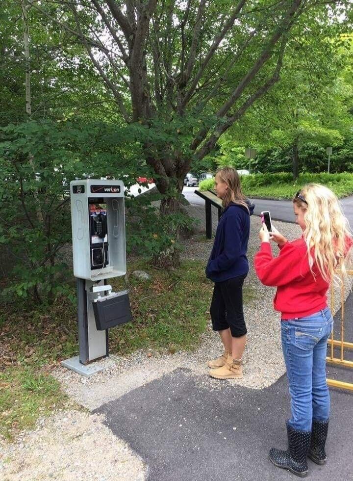 Two people stand near a tall public calling or vending machine in an outdoor area, a person in a red hoodie is filming with a smartphone while the other person waits nearby.