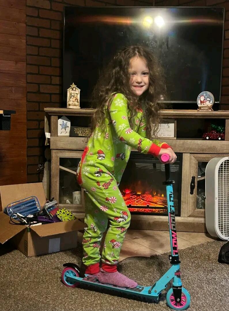 A young girl with curly hair, wearing green Christmas-themed pajamas, stands on a blue and pink scooter in a living room. In the background, there's a TV, a fireplace with artificial flames, and Christmas decorations including signs that say 'MERRY CHRISTMAS' and 'LET IT SNOW'. The scooter has 'FLOW' and 'MAD' visible on it.