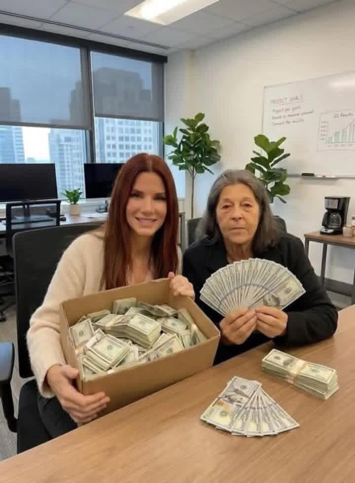 Two women in an office, one holding a box full of money, the other holding a fan of dollar bills. Stacks of money are also on the table.