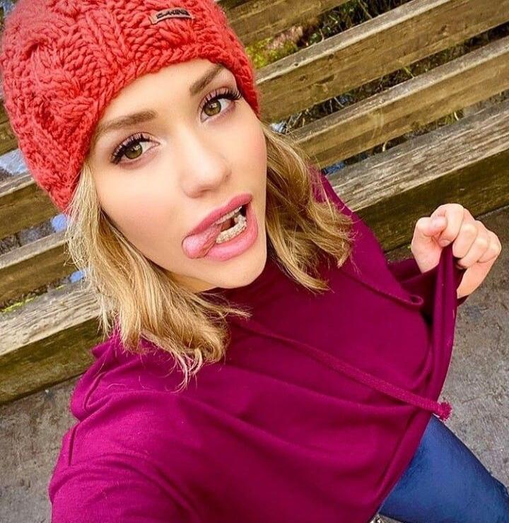 A young woman wearing a red knitted hat and a burgundy hoodie, sticking her tongue out and showing braces, making a playful face. She is holding up her hoodie slightly with one hand.