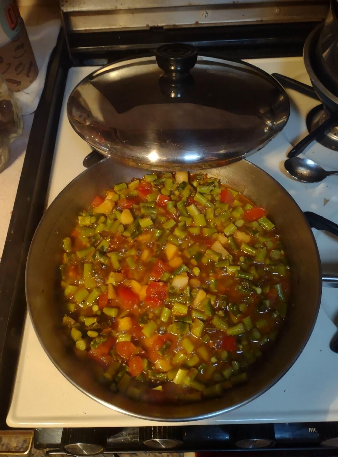 A pan of diced vegetables (green, red, orange, yellow) cooking on a stove with a glass lid nearby.