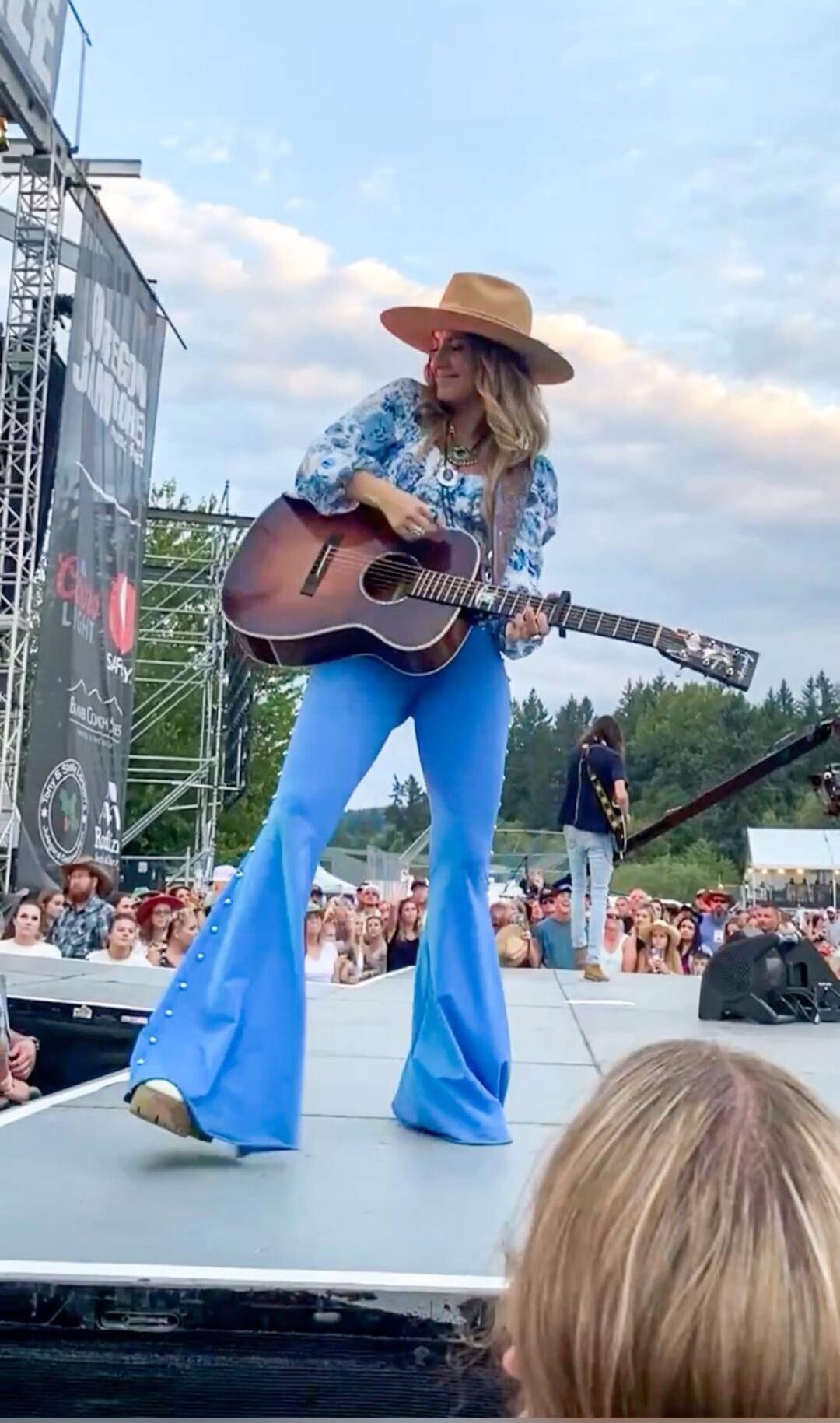 A female musician wearing a wide-brimmed hat performing on stage with an acoustic guitar at an outdoor concert; crowd in the audience.