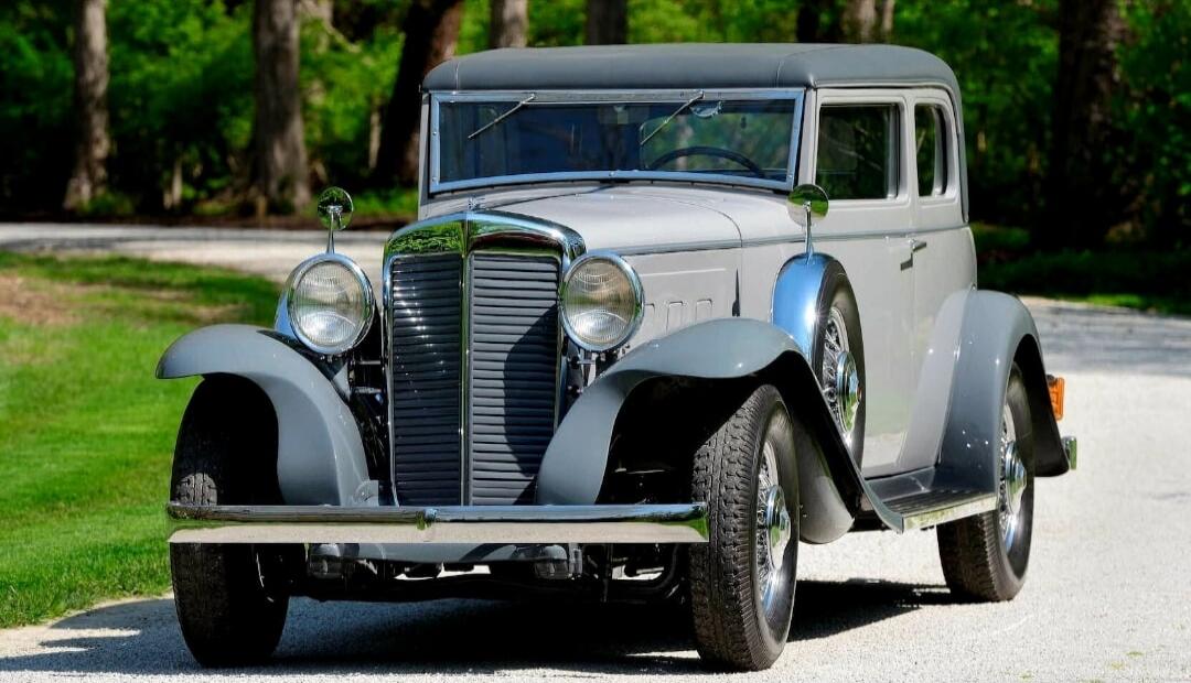 A vintage grey car with chrome accents is parked on a gravel path, surrounded by green grass and trees in the background.