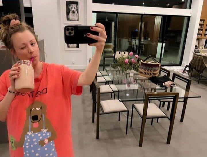 A woman in a coral t-shirt with a cartoon dog design and reversed text 'Do Lisa' is taking a selfie, winking and drinking from a jar with a pink straw. In the background, there is a modern dining area with a glass table and chairs, and a framed picture of a dog on the wall.