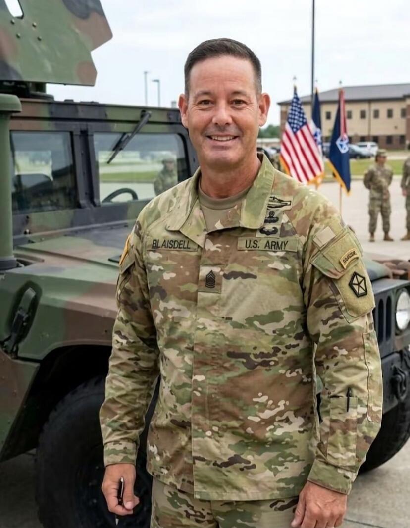 A man in a U.S. Army uniform, with the name BLAISDELL and rank insignia, stands next to a military vehicle. U.S. flags are visible in the background.