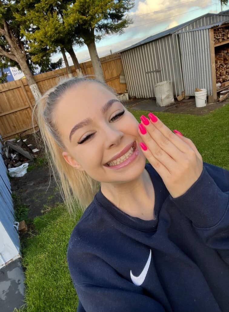 A young woman with blonde hair in a ponytail, wearing a dark blue Nike sweatshirt, smiles and winks at the camera. She has bright pink nail polish on her fingers, which she holds near her face. In the background, there's a green lawn, a wooden fence, trees, and a metal shed with stacked firewood. A partially visible sign reads 'for pride'.