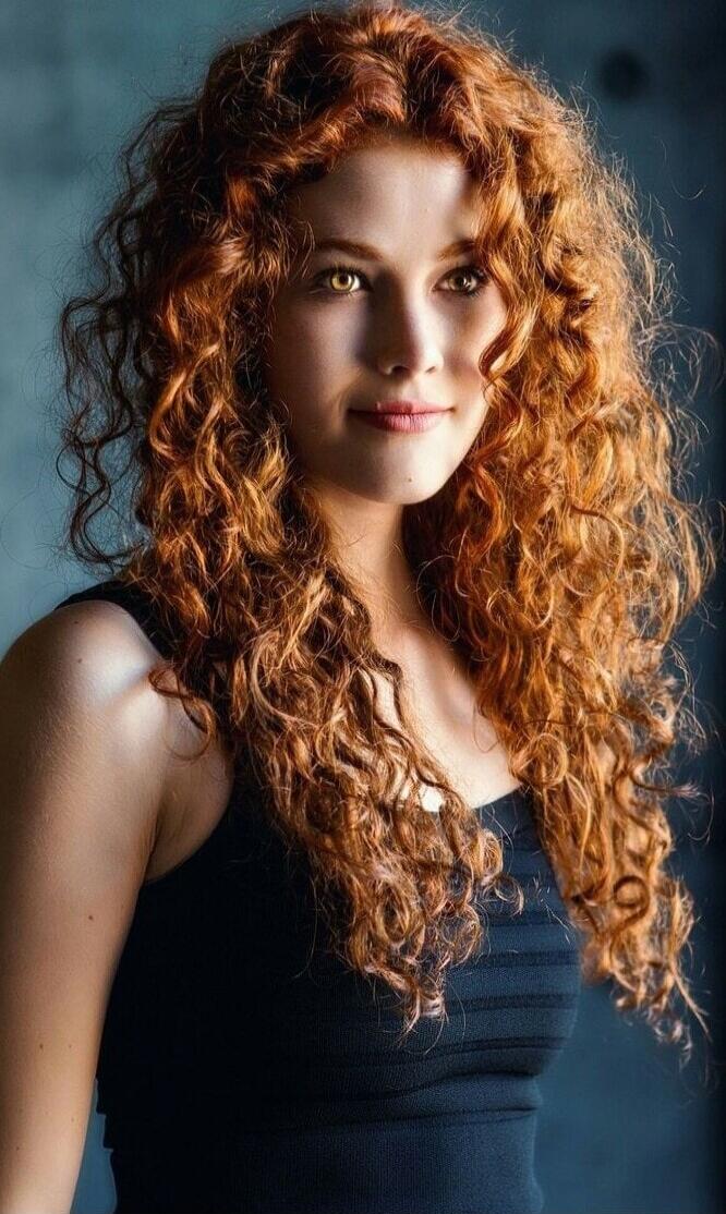 A portrait of a young woman with long, curly red hair wearing a black top.