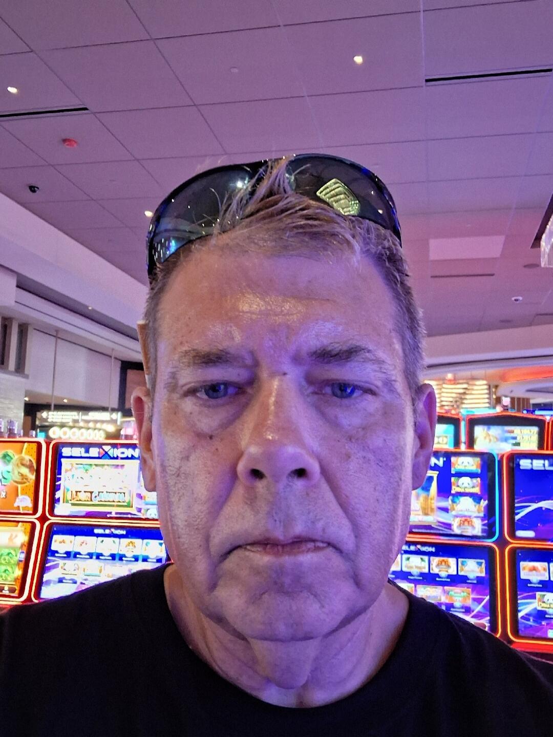 A man takes a selfie in a casino, with slot machines in the background. He has sunglasses on his head and a neutral expression. Text visible on machines includes 'SELEXION' and 'GOODWIN'.