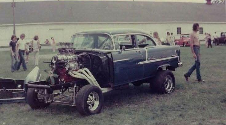 A vintage dark blue Chevrolet gasser or drag car with its front end removed, showcasing a large, powerful engine with multiple carburetors and long exhaust headers. The car has large rear tires and smaller front tires. Several people are in the background, and a shirtless man in jeans walks away in the foreground. The scene is an outdoor car event 