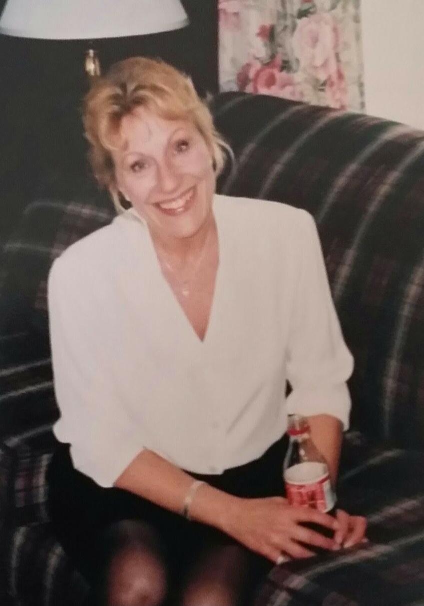 A woman sits on a plaid couch, wearing a white blouse and dark skirt, smiling and holding a bottle (appears to be a soda bottle).