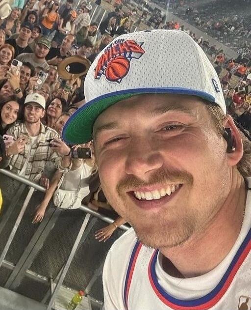 A man wearing a white New York Knicks baseball cap and a basketball jersey is taking a selfie. He has a mustache and is smiling broadly. Behind him, a large crowd of people can be seen, suggesting he is at a public event like a concert or a sports game. The text 'NEW YORK' and a basketball logo are visible on his cap.