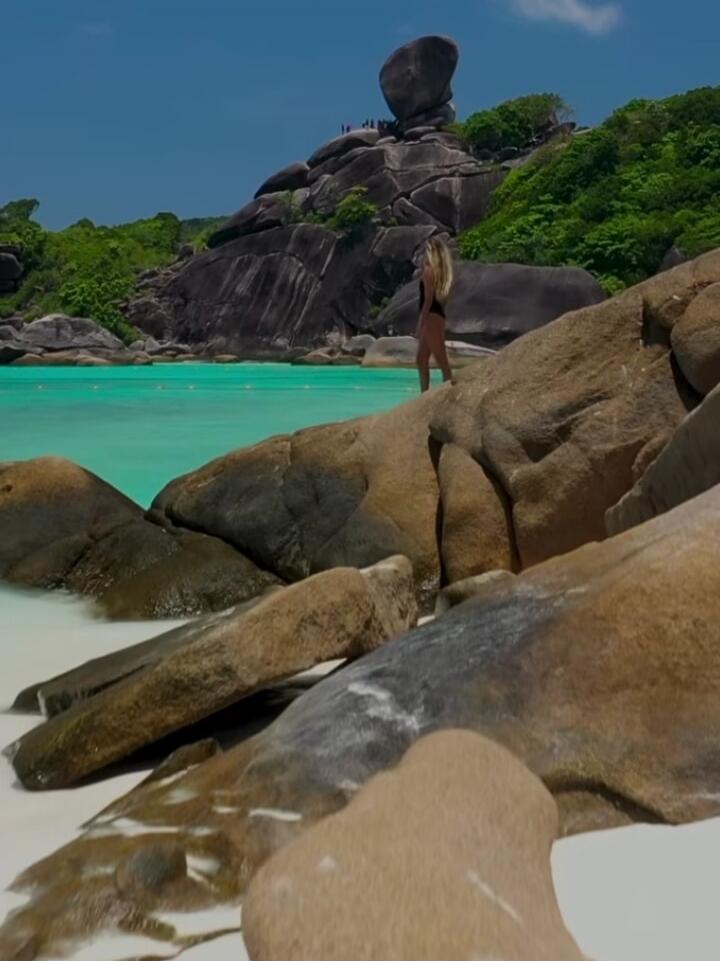 A person in a swimsuit stands on large rocks by a turquoise tropical beach with green trees in the background.
