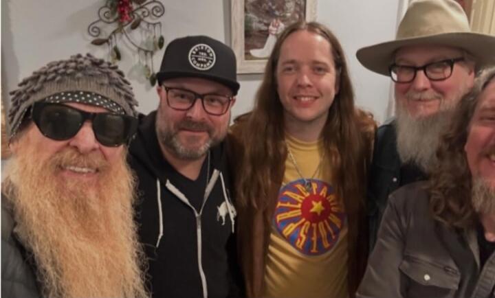 Five men posing for a group photo; one wearing sunglasses and a beanie, another with a sun logo shirt.