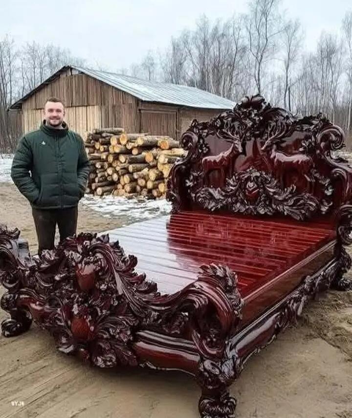 A man standing next to an ornate, dark wood carved bed in a rural yard with a woodpile and a shed.