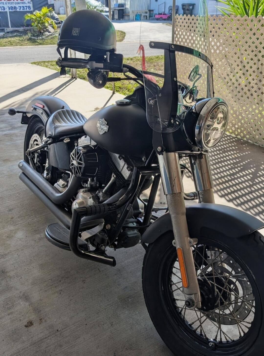Matte black Harley-Davidson cruiser motorcycle with windshield and helmet resting on the handlebars, parked in a driveway.
