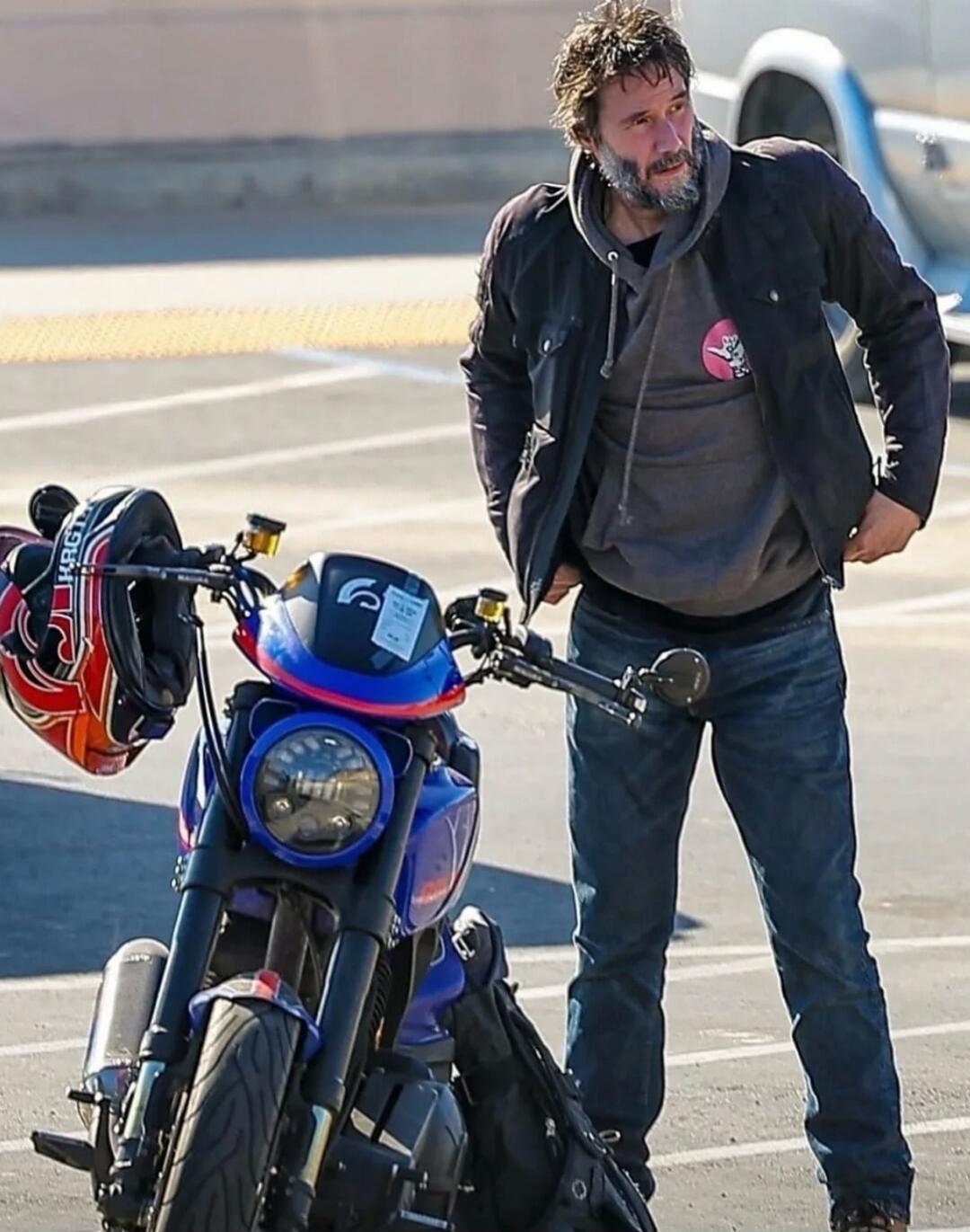 Bearded man standing next to a blue motorcycle in a parking lot.