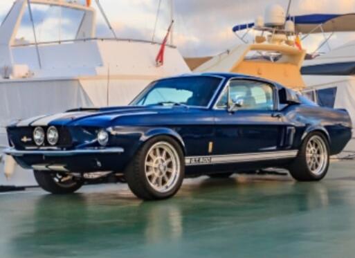 A classic blue Ford Mustang Shelby GT500 parked on a dock with yachts in the background. The car features white racing stripes and the 'GT 500' emblem on its side.