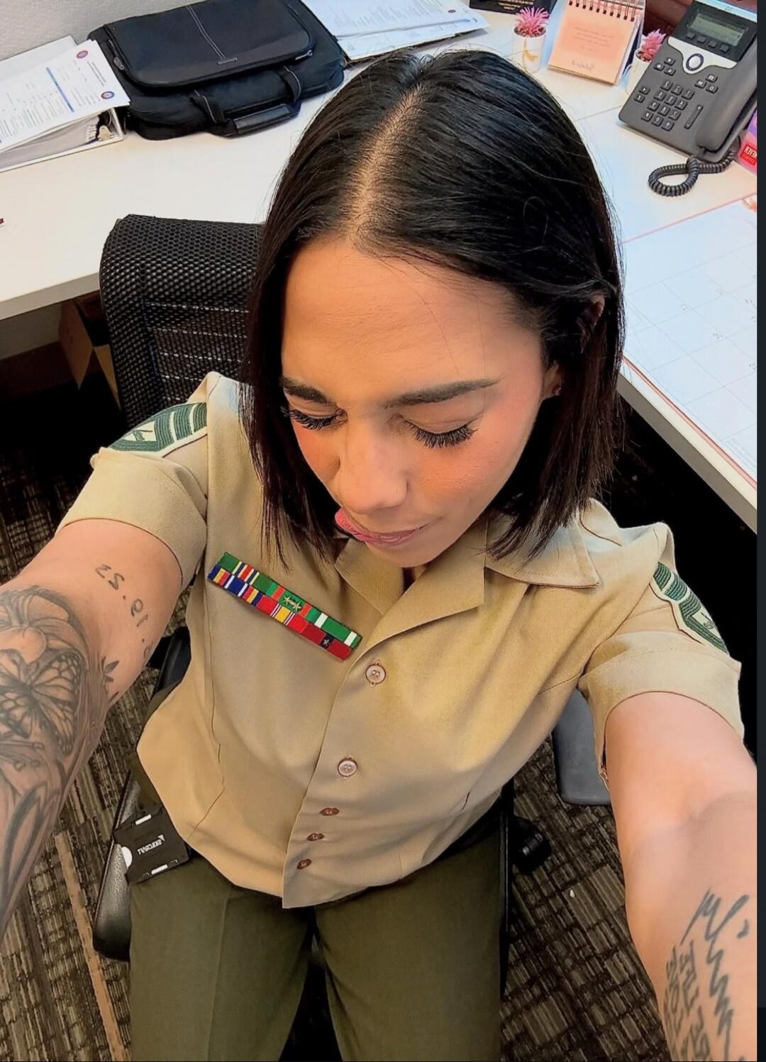 A woman in a military uniform, possibly a Marine, is taking a selfie at an office desk. She has ribbons on her chest and tattoos on her arms. Her head is tilted down, and her tongue is slightly out. Text visible on her left arm tattoo reads '25.er.' A device clipped to her pants has 'PROXIM' (reversed) written on it.