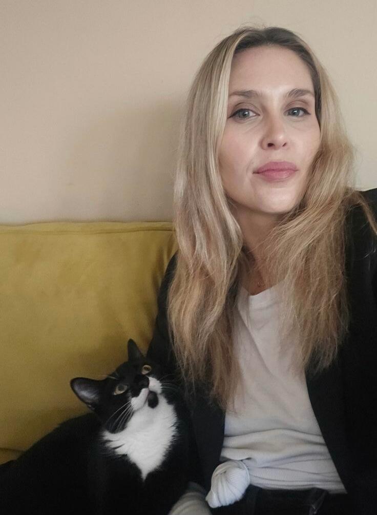 A woman with long blonde hair and a black and white cat are sitting on a yellow couch.