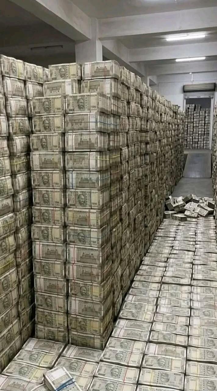 A large room filled with massive stacks of Indian 500 Rupee banknotes. The currency notes are bundled and piled high against the walls and spread across the floor, indicating an immense amount of money.