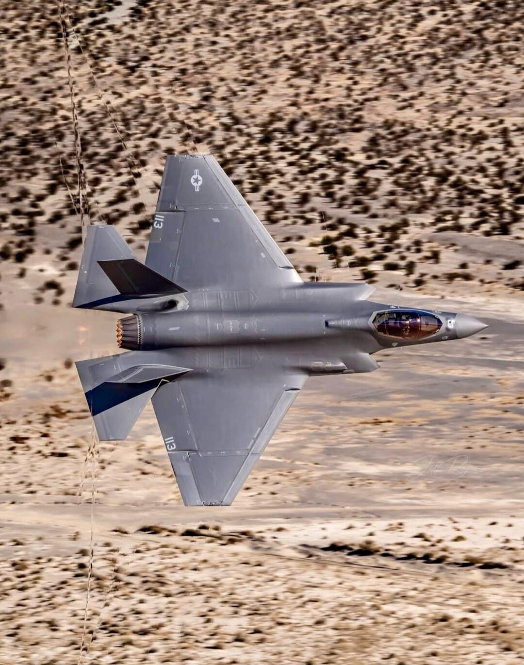 An F-35 fighter jet flying low over a desert landscape, with its afterburner glowing.