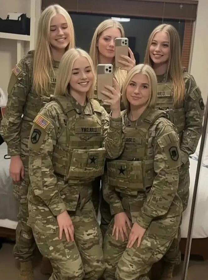 Five young blonde women in military camouflage uniforms and tactical vests are smiling and taking a selfie indoors. They are wearing patches with the American flag and what appears to be unit designations like 'YHO AR.U' and 'AFR.U' (likely reversed text). Black stars are visible on their vests.