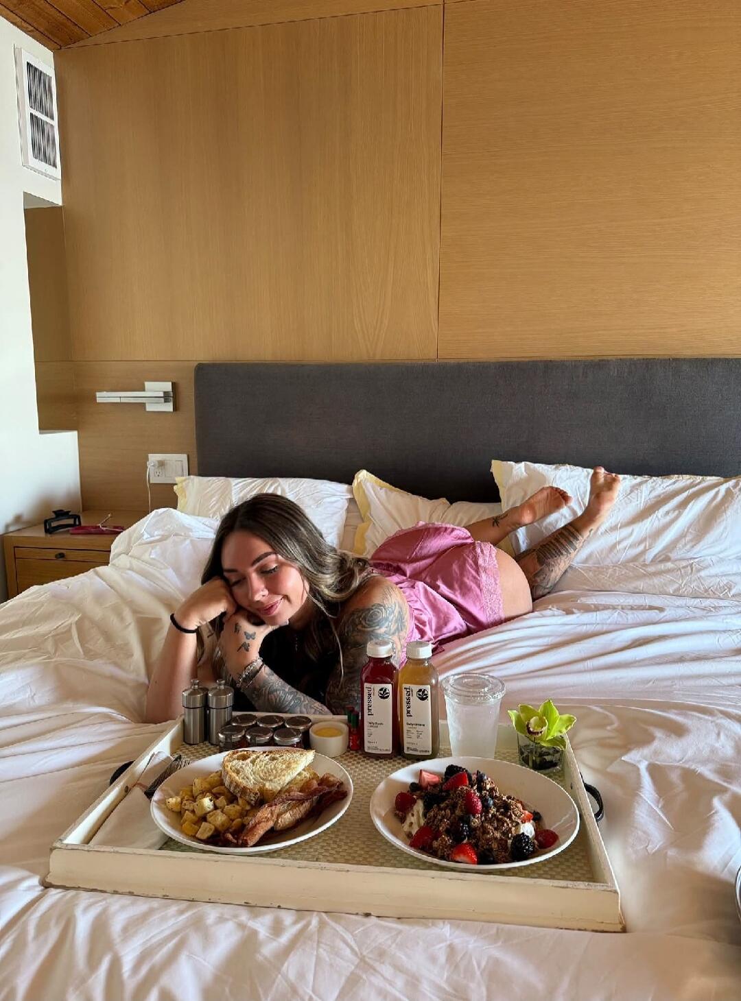 A woman is lying on a bed, smiling, with a breakfast tray in front of her. The tray contains plates with food like bacon, potatoes, toast, and a bowl of berries and granola. There are also two bottles labeled 'pressed!' and a glass of water. She is wearing pink sleepwear. The setting appears to be a hotel room.